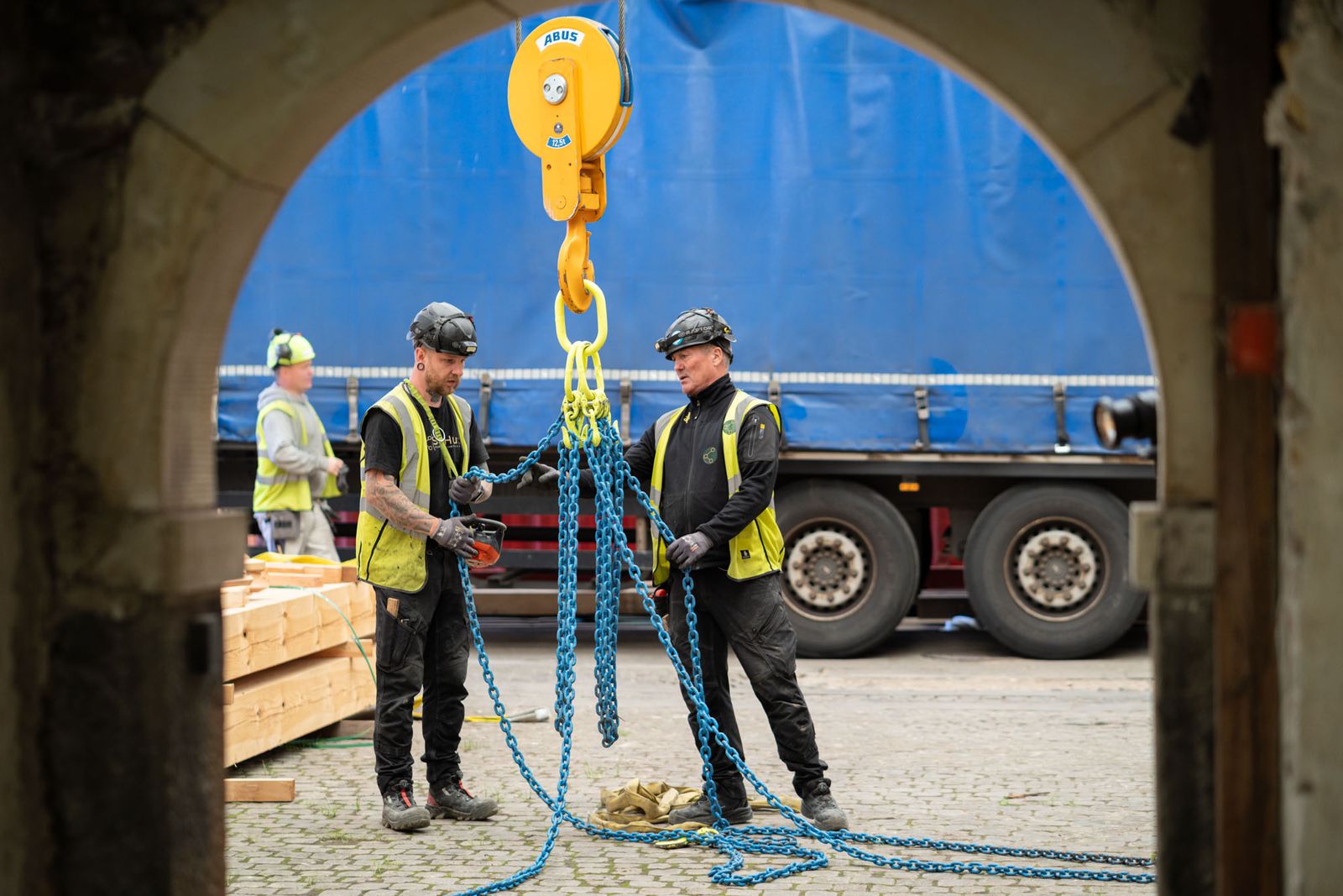 Tre pladsfolk fra SiteHub gør en kædestrop klar under en portbue med en lastbil bag sig