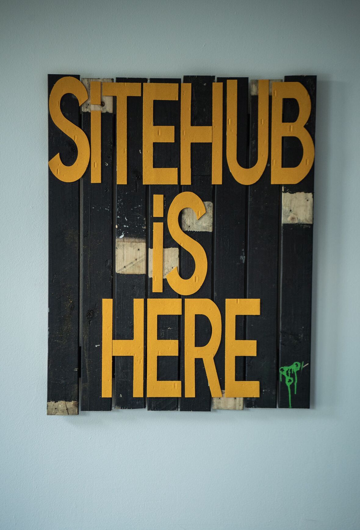 Skilt af sortmalede brædder på en væg med gule bogstaver: SITEHUB IS HERE