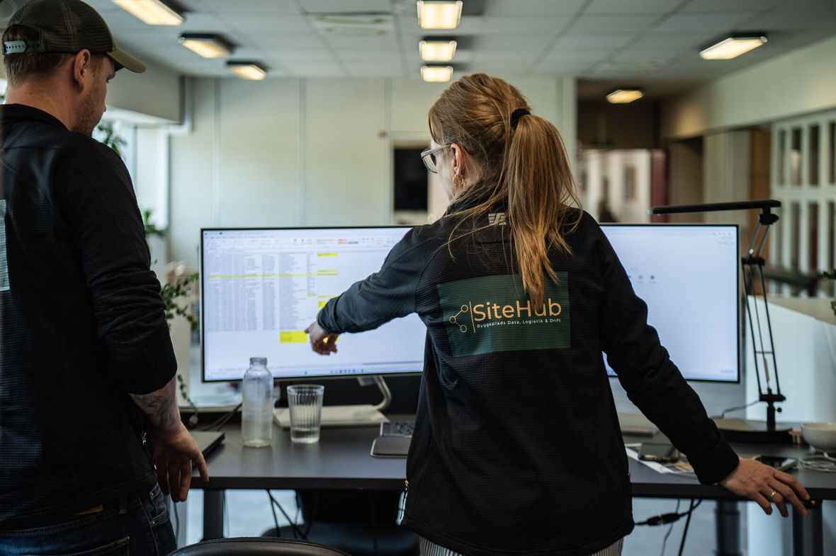 SiteHub-medarbejder peger på et regneark på en bred skærm, mens en kollega ser med, på kontoret