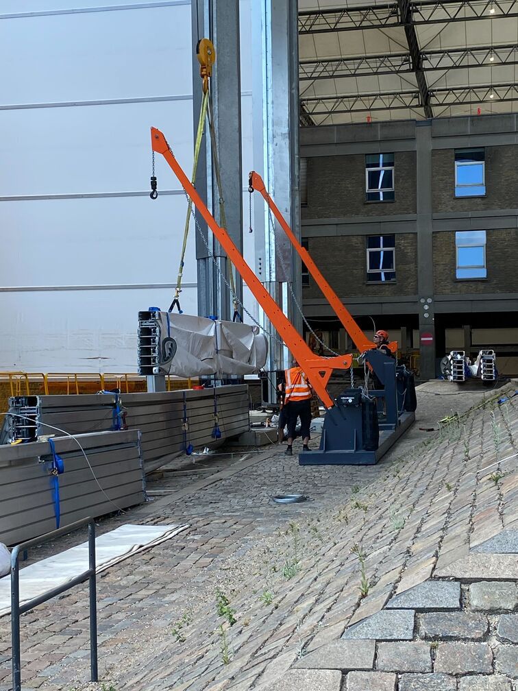 Orange løftebom hejser et indpakket byggeelement på et brostensbelagt gårdareal