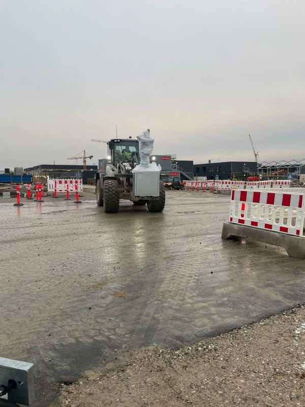 Gummihjulslæsser kører over en våd betonflade mellem afspærringer med byggekraner i baggrunden